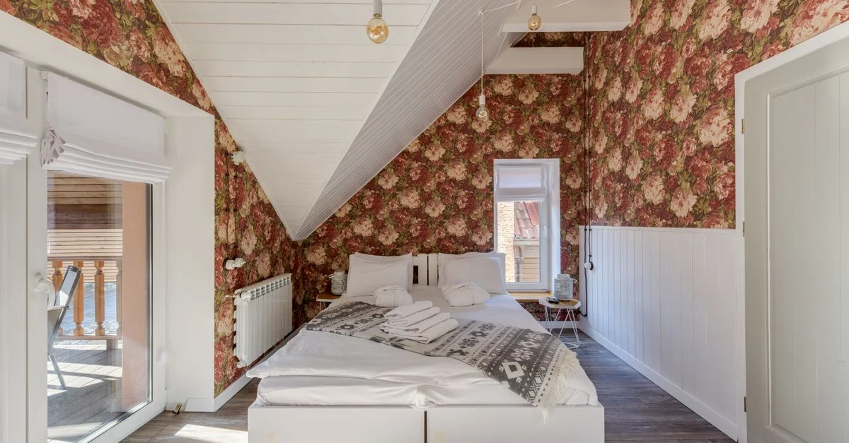 Chambre de mansarde lumineuse avec papier peint floral, lit et lumière naturelle provenant de fenêtres en verre.